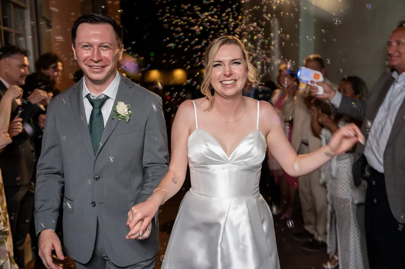 Tim in a gray suit and Sarah in a white slip dress hold hands and smile while walking through a shower of confetti