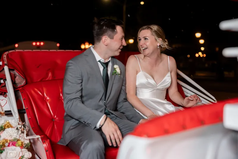 Sarah and Tim sit together in a red vintage car at The White Room, smiling at each other during their reception.