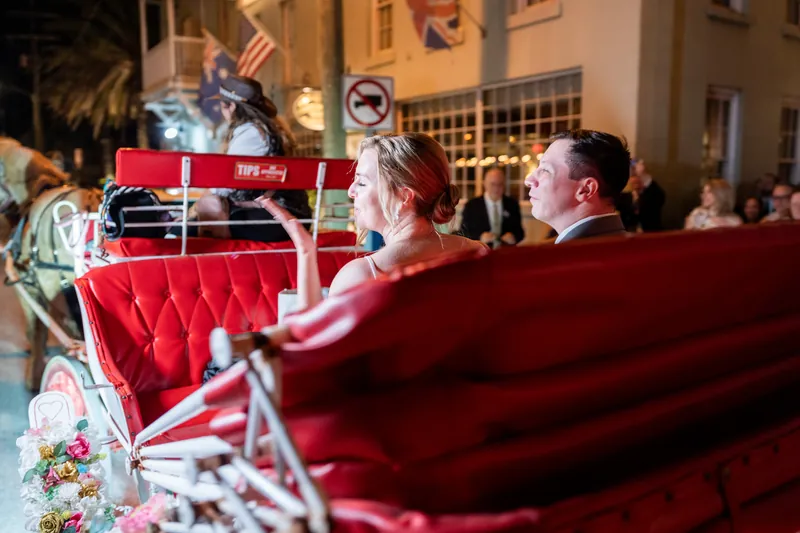 Sarah and Tim ride in a red vintage car decorated with flowers, waving as they depart The White Room.