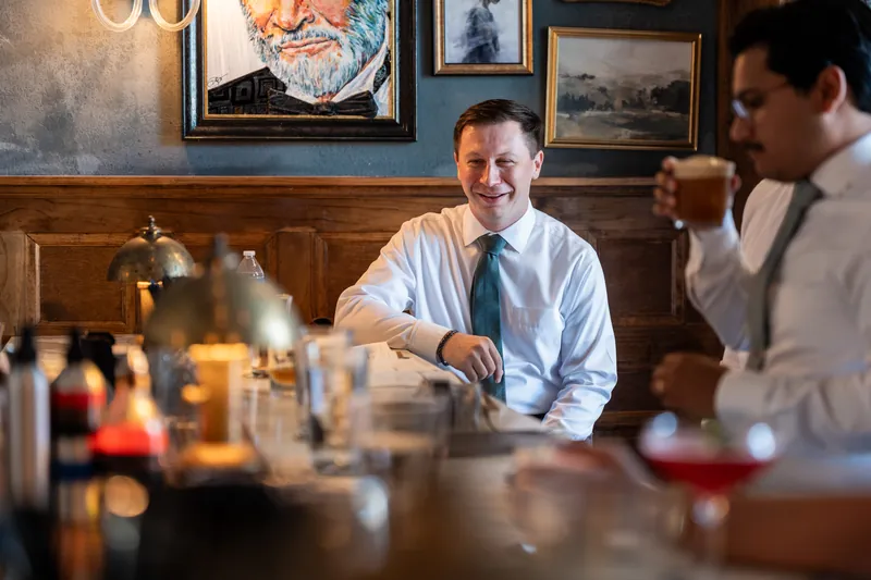 Tim sits at a wooden bar wearing a light blue shirt and teal tie, chatting with a guest.