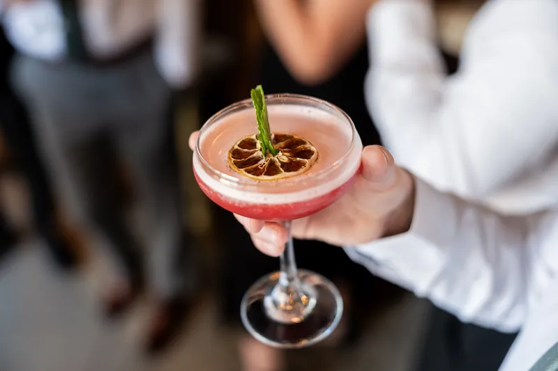 Someone holds a pink cocktail garnished with a rosemary sprig and dried citrus at The White Room.