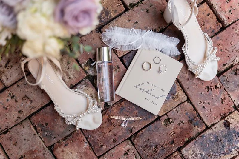 Wedding rings, white shoes, perfume, and flowers arranged on brick pavement at The White Room.