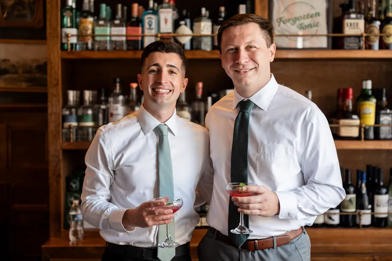 Tim and a groomsman smile while holding drinks in front of a wood-paneled bar stocked with bottles.