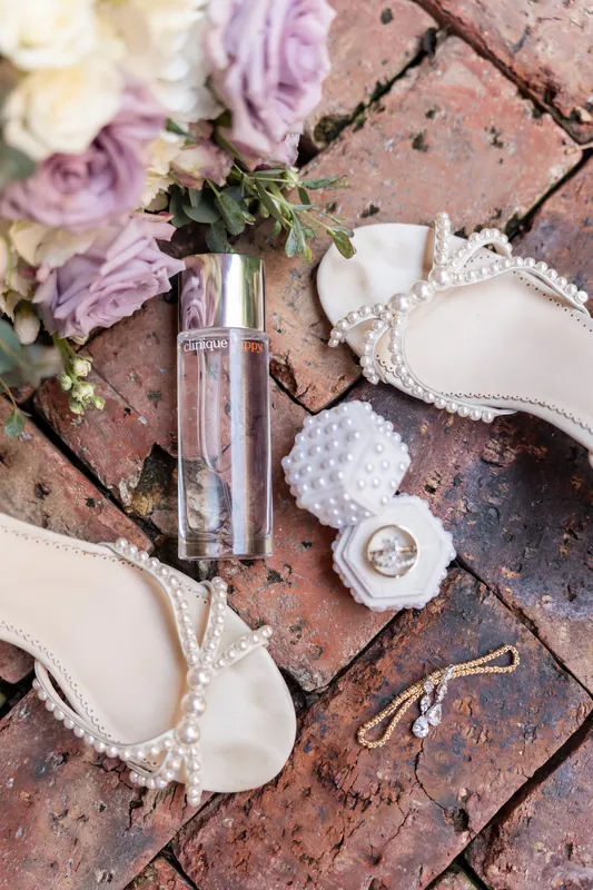 White heeled shoes, rings, and a clear fragrance bottle sit among purple and white florals on brick.