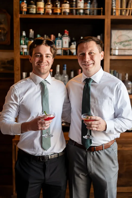 Tim and his groomsman stand side-by-side in white dress shirts and ties, each holding a cocktail.