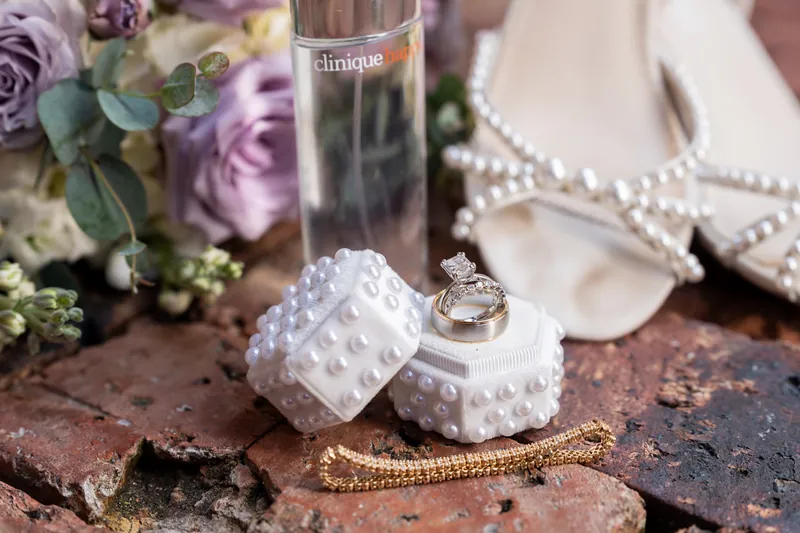 Wedding bands and a pearl bracelet rest on white textured boxes next to purple flowers and perfume.