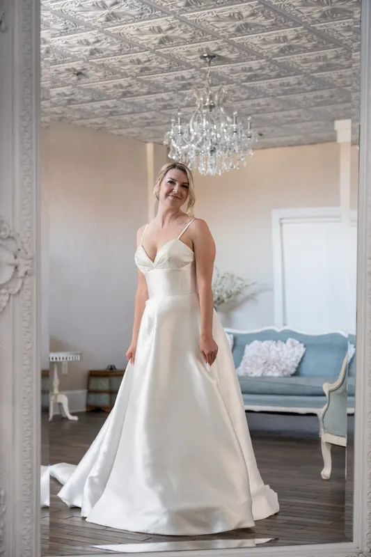 Sarah stands alone in her flowing white satin gown beneath a crystal chandelier at The White Room.