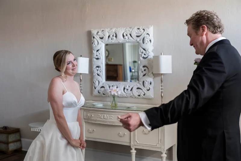 Tim gestures while speaking with Sarah, who watches him in her white gown near a decorative mirror.