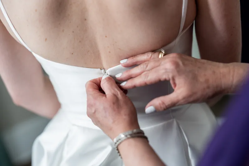 Hands adjust the back of Sarah's white dress, fastening details at The White Room before the ceremony.