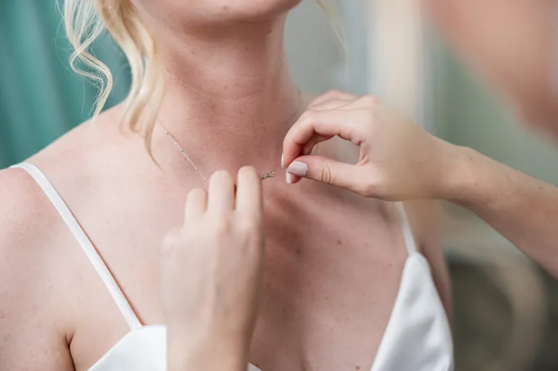 Someone fastens a delicate necklace around Sarah's neck at The White Room, completing her bridal look.