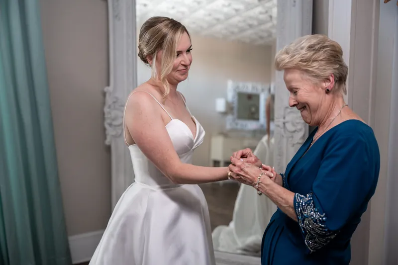 Sarah extends her arm to receive help with jewelry from a woman in a navy cardigan.
