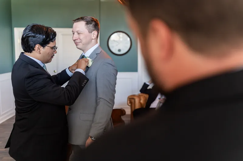 A man in a dark suit helps Tim with his tie as other guests look on in the background.