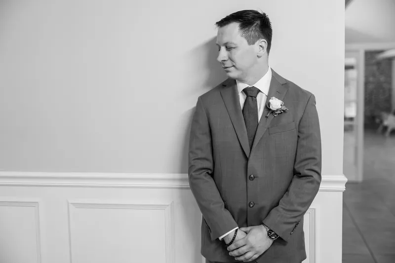 Tim stands against a white paneled wall in a gray suit with a boutonniere, looking toward the side.