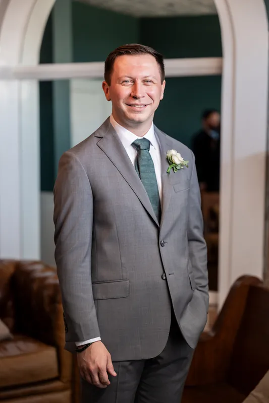 Tim stands in a gray suit with a boutonniere, smiling directly at the camera in the venue's interior.