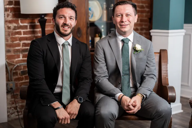 Tim and a groomsman sit together on a brick-exposed bench, both in suits with boutonnieres.