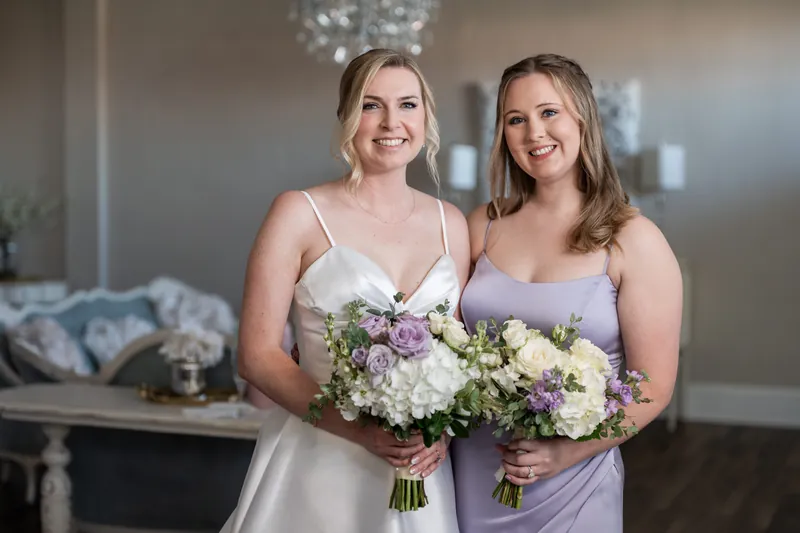 Sarah and a bridesmaid stand together holding white and purple bouquets, both beaming.