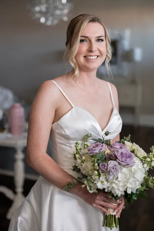 Sarah holds a white and lavender bridal bouquet at The White Room, smiling at the camera in her sleeveless ivory gown.