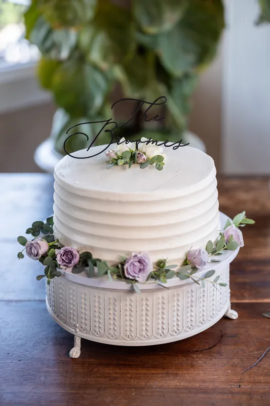 A two-tier white wedding cake topped with "The Grooms" sign and decorated with purple and white florals sits on a table.