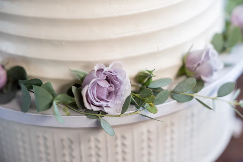 Purple roses and eucalyptus garland drape across the tiered wedding cake's white textured base.