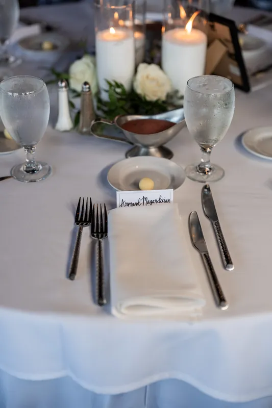 An elegant place setting at The White Room features white linens, candles, and a handwritten name card beside a white rose.