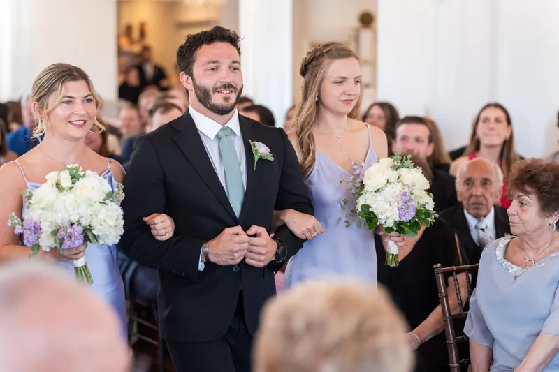 Tim walks down the aisle flanked by two women holding white and purple bouquets at Villa Blanca.