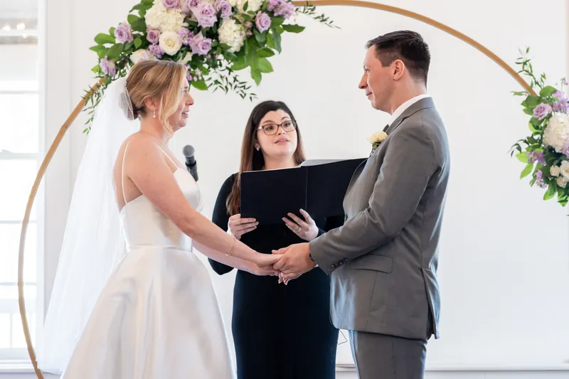 An officiant holds a black folder as Sarah and Tim face each other beneath a curved floral arch during their vows.