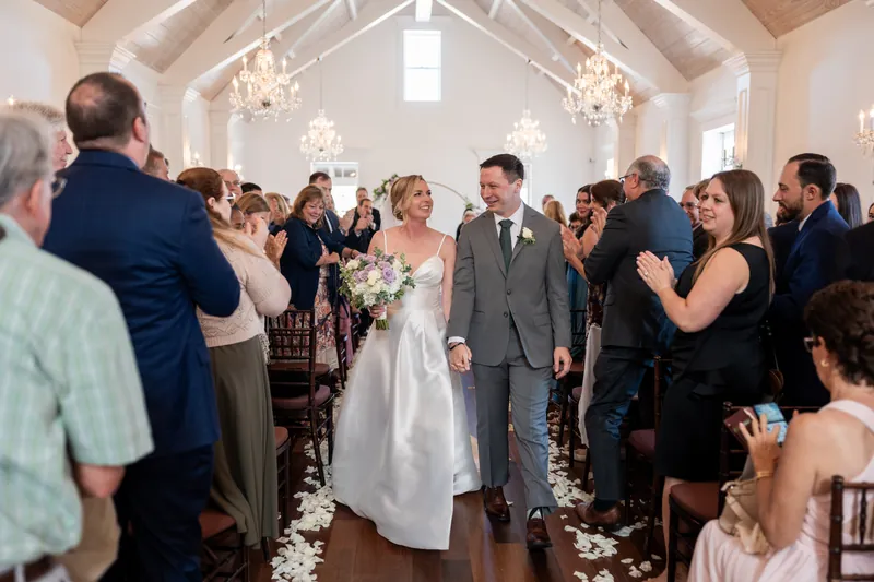 Sarah and Tim walk through a flower-lined aisle as wedding guests applaud in the bright, vaulted chapel.