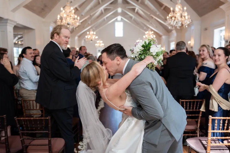 Tim dips Sarah in a kiss as guests cheer around them in the elegant white wedding space.