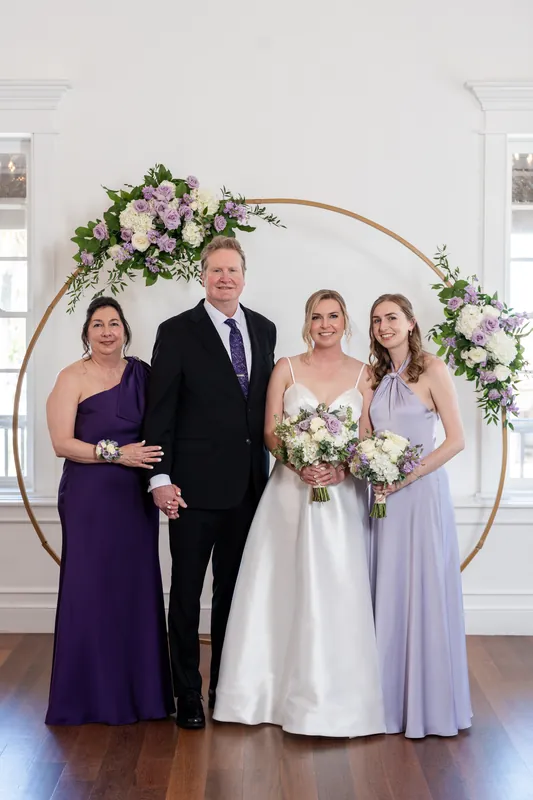 Sarah and Tim stand with three attendants beneath a floral-draped arch at Villa Blanca, St. Augustine, wearing formal attire