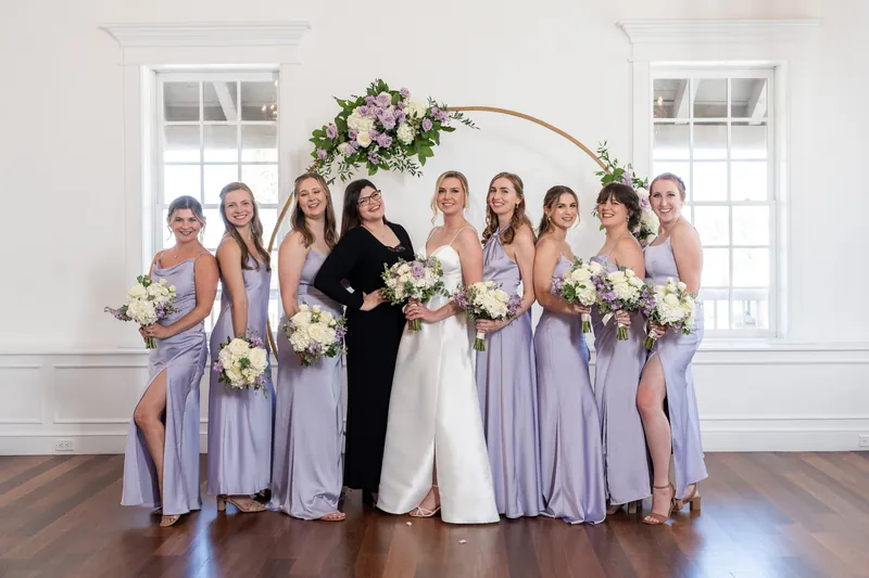 Sarah and Tim pose with nine attendants in lavender and charcoal suits beneath the arch, all holding white and blush