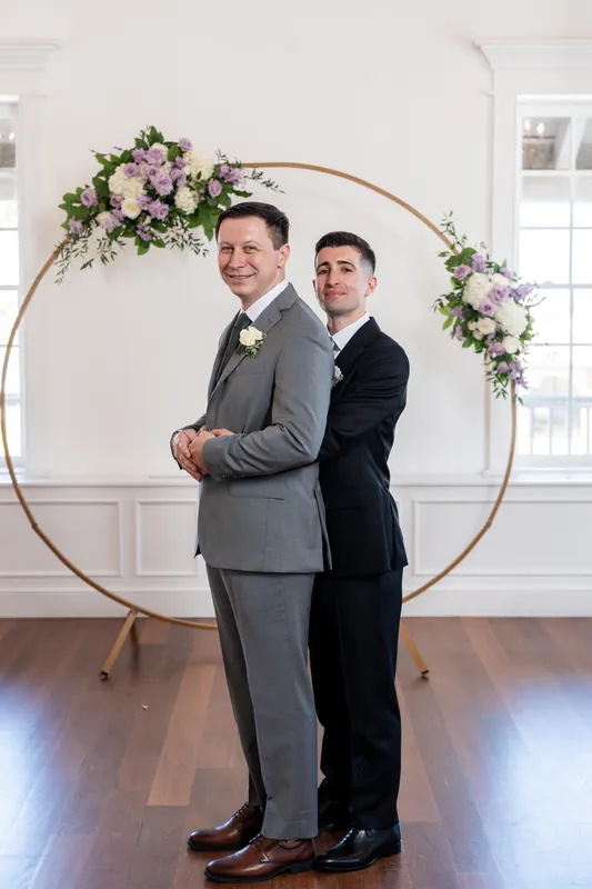 Tim and his partner embrace while standing beneath the floral arch, both smiling in gray and navy suits.