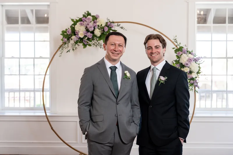 Two groomsmen stand shoulder-to-shoulder beneath the arch, both wearing boutonnières and formal suits.