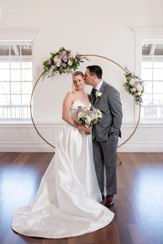 Sarah and Tim stand together beneath the arch; Sarah wears a white gown with a long train while Tim wears a gray suit, both