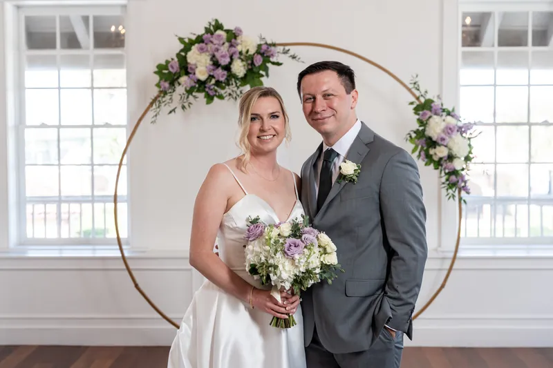 Sarah and Tim stand together under a floral arch at Villa Blanca, Sarah holding a white and purple bouquet.