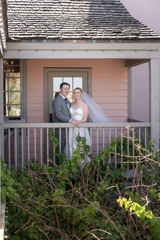 Sarah and Tim pose on a cottage porch railing with ivy and flowering vines below.