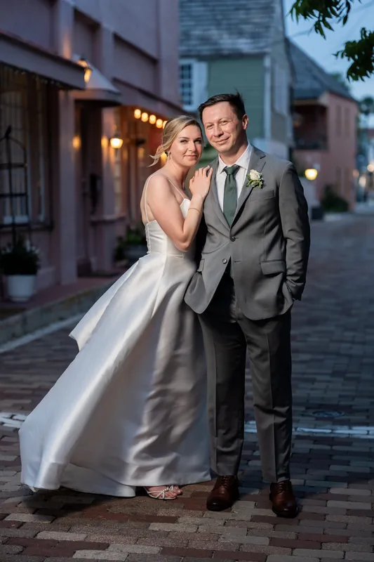 Sarah leans against Tim on a brick street at twilight, her white gown's train swept across the pavement, palm trees framing