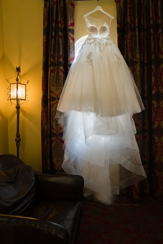 A tiered wedding dress hangs on a wooden frame against golden walls and burgundy curtains at Casa Monica Resort & Spa.
