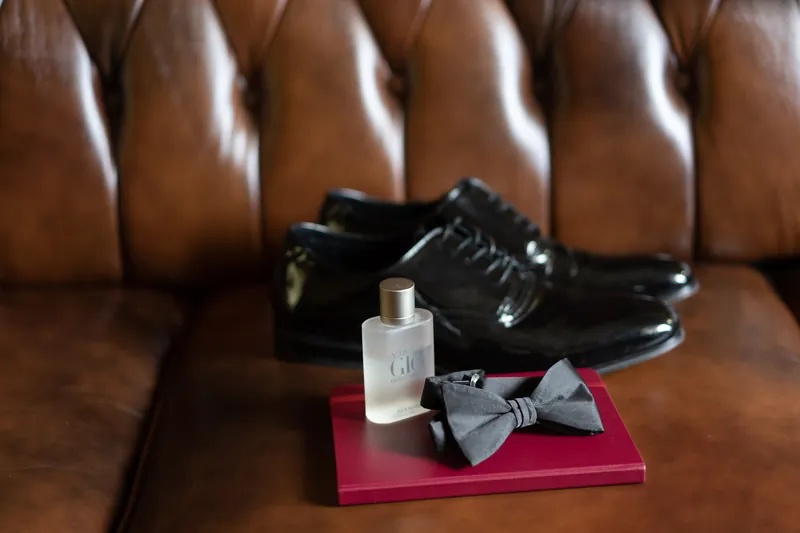 Polished black dress shoes and a cologne bottle sit on a pink platform atop a brown leather bench.