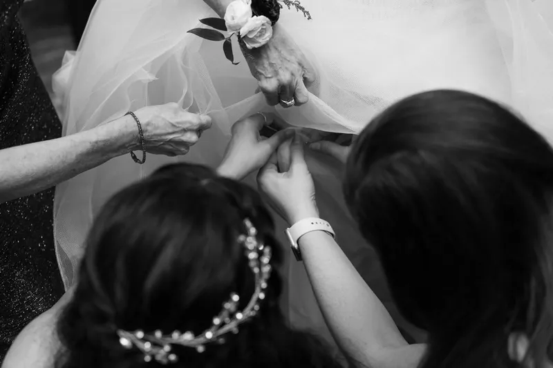 Attendants help adjust Simone's wedding dress and bouquet in a close-up black-and-white moment.