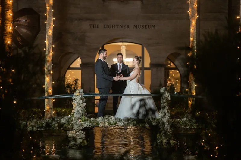 Michael and Simone kiss on an arched bridge flanked by lit columns and white florals.