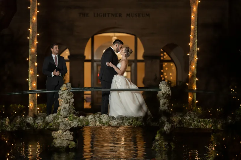 Simone and Michael kiss beneath an illuminated archway with greenery and water reflections below.