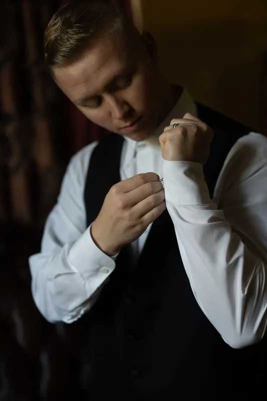 Michael fastens his white dress shirt cufflinks while wearing a black vest in soft, dramatic lighting.