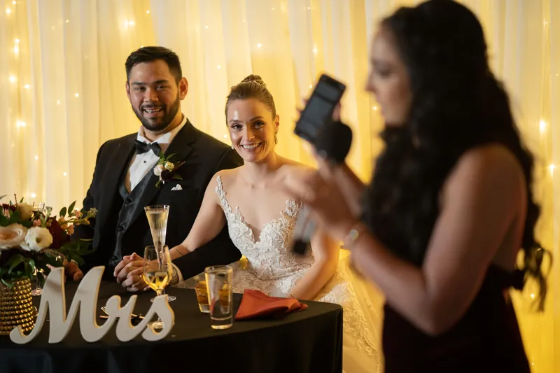 Simone laughs while Michael watches a guest speak at the reception table, a gold "Mrs" sign visible nearby.