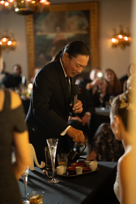 Michael holds the microphone and gestures while speaking to guests gathered around the reception hall during his toast.
