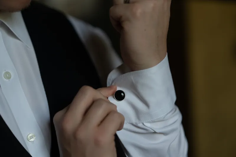 Hands adjust a dark cufflink on Michael's crisp white shirt sleeve during final grooming preparations.