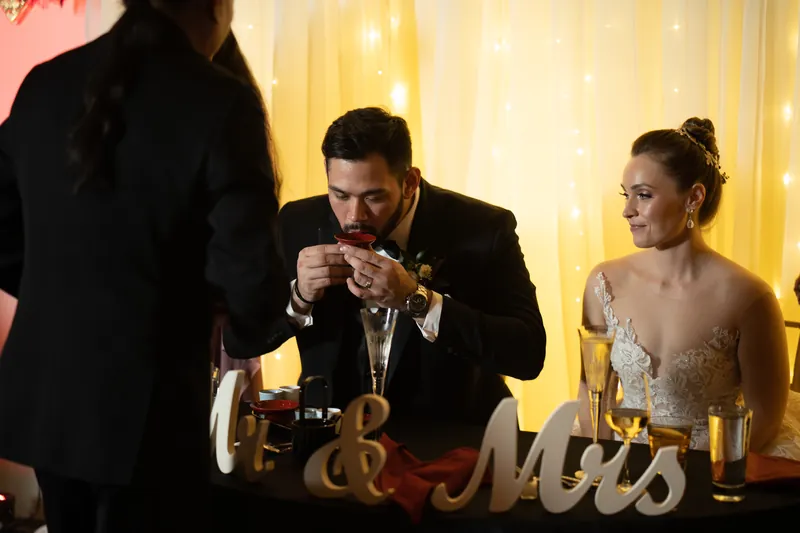 Michael and Simone sit together at their reception table, sharing a quiet moment as a guest stands nearby.