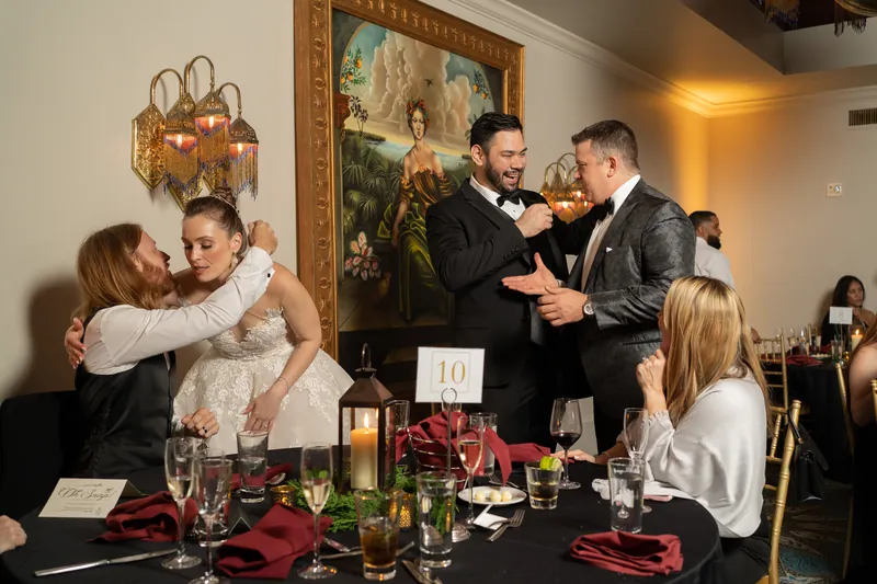 Michael adjusts his bowtie while a groomsman helps as family members embrace at the reception table.