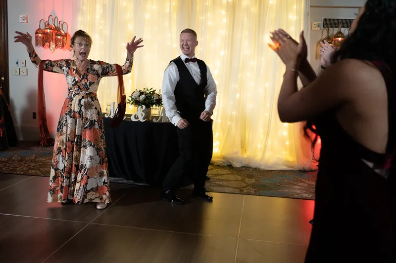 An elderly woman with arms raised dances on the lit dance floor while Michael watches and guests clap along.