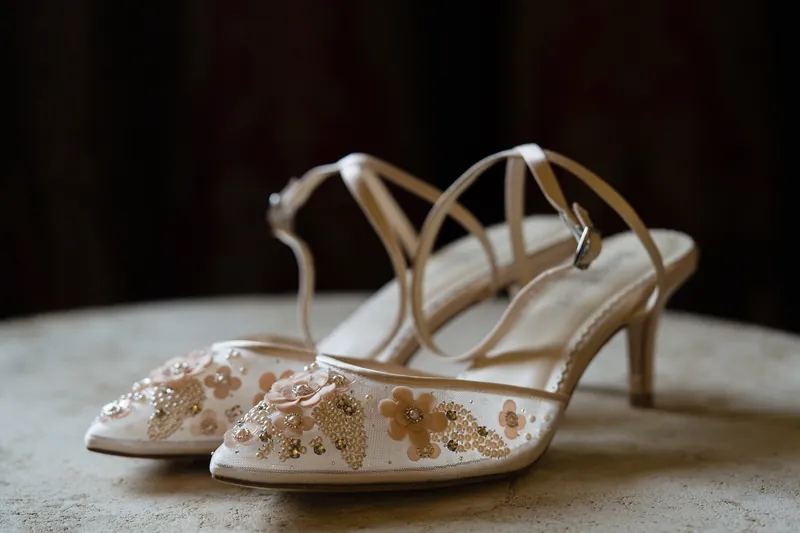 Ivory kitten heels with beaded floral embellishment sit on a pale surface against a dark backdrop.