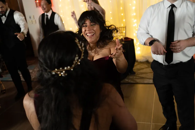 A woman in a black dress laughs and dances with an animated expression while men in formal wear surround the dance floor.
